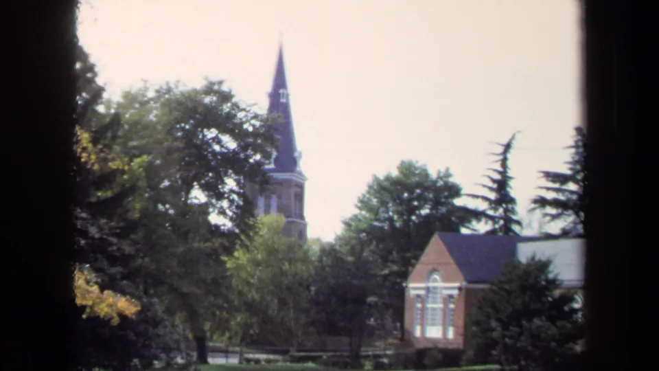 1970 8mm Washington, DC campus view with a steeple, brick building, shrubs, and autumn trees beyond dark window framing.