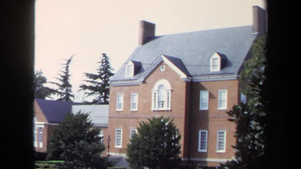 Washington, DC 1970 8mm frame centered on a large brick building with white-trimmed windows, roof dormers, shrubs, and trees.