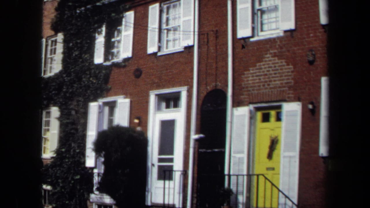 1970 Washington, DC 8mm frame showing brick row houses with white shutters, iron railings, ivy, and a yellow front door.