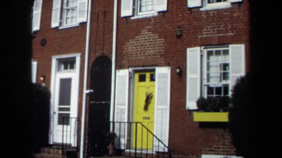 1970 Washington, DC home-movie frame showing a brick townhouse entry with a yellow door, white shutters, iron railing, and window box.