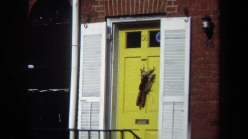 Close 1970 Washington, DC exterior frame of a yellow townhouse door with a wreath, white shutters, brick wall, and black railing.