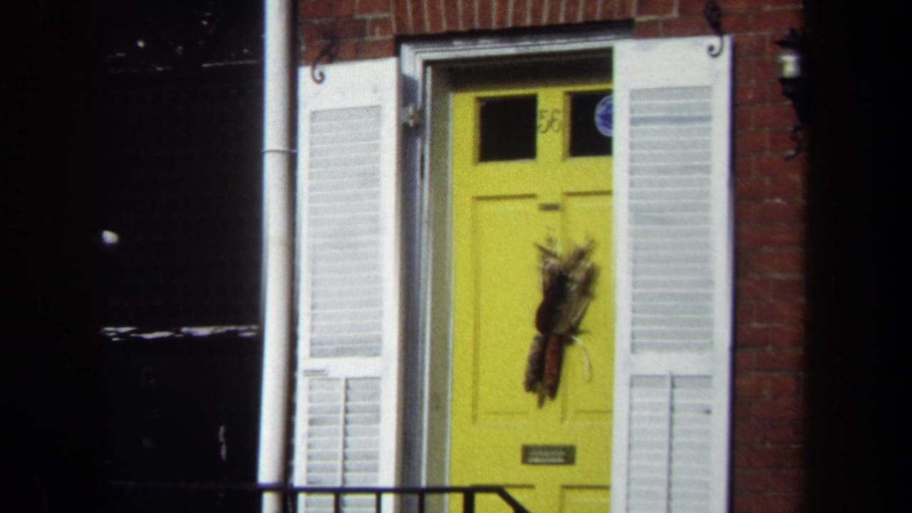 1970 Washington, DC home-movie frame showing a yellow front door, white shutters, brick facade, and dark foreground shadows.