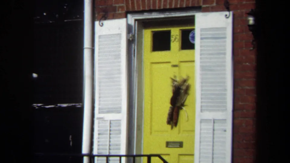 1970 Washington, DC home-movie frame showing a yellow front door, white shutters, brick facade, and dark foreground shadows.