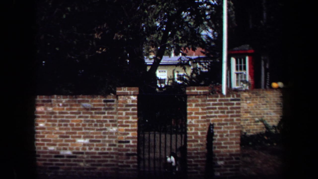 1970 Washington, DC home-movie frame with a brick boundary wall, black gate, trees, and a partially visible house beyond.