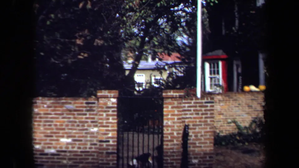 1970 Washington, DC home-movie frame showing a brick wall, dark metal gate, leafy branches, and part of a residence behind it.