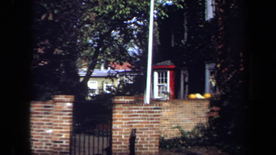 1970 Washington, DC home-movie frame with a brick wall and gate in the foreground and a shaded residential exterior beyond.