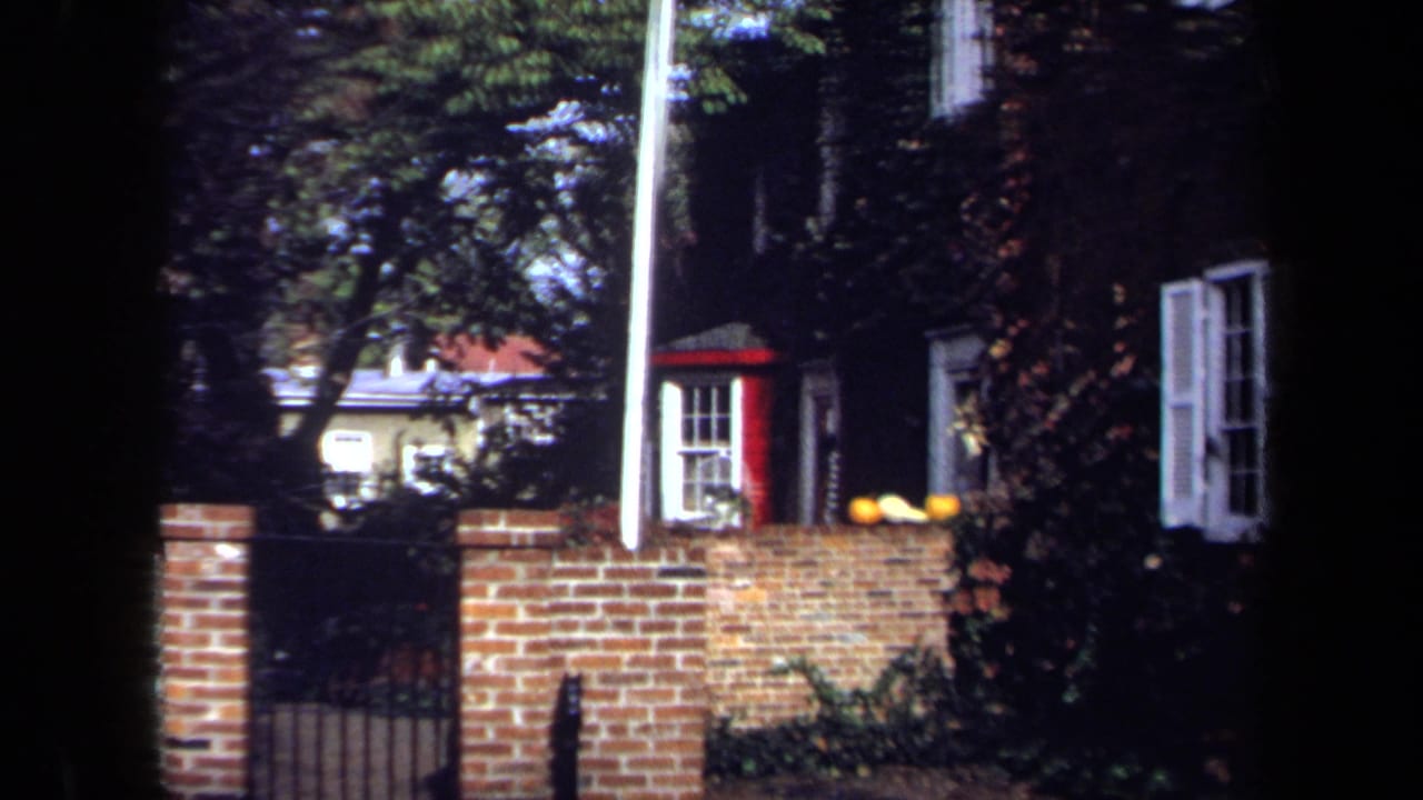 1970 Washington, DC 8mm view of a brick townhouse partly framed by trees, a brick wall, white shutters, and a red-trimmed bay window.