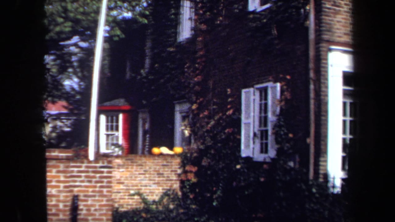 1970 Washington, DC home-movie frame showing white-shuttered windows, ivy on brick, a red-trimmed bay window, and the foreground wall.