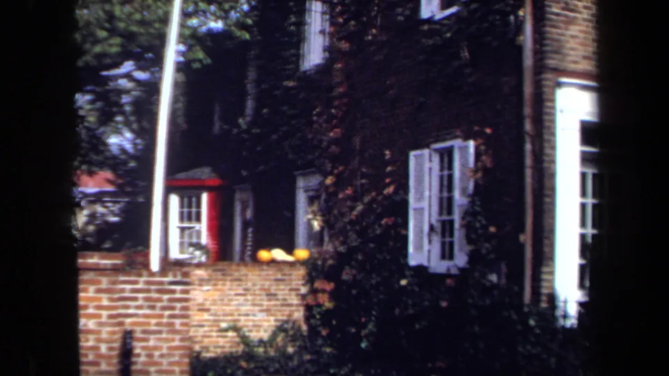 1970 Washington, DC home-movie frame showing white-shuttered windows, ivy on brick, a red-trimmed bay window, and the foreground wall.