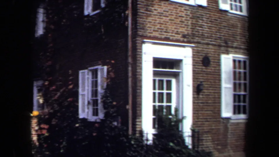 1970 Washington, DC 8mm frame of a brick townhouse entrance and windows, partly obscured by dark foreground trees and climbing ivy.