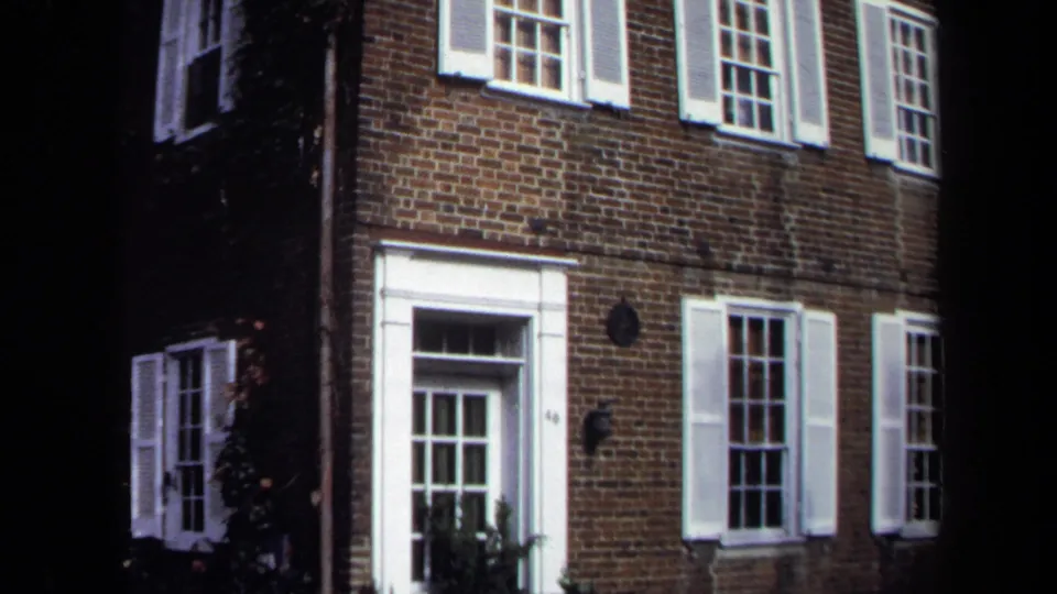 1970 Washington, DC home-movie frame showing a brick townhouse entrance, white shutters, multi-pane windows, ivy, and ironwork near the door.