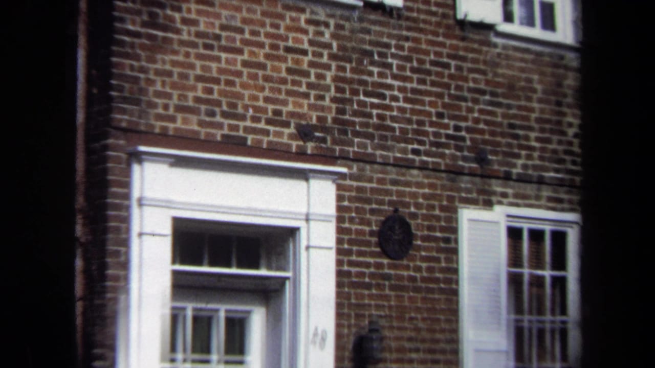 1970 Washington, DC home-movie frame showing a brick facade with white-trimmed entry, window shutters, and a dark wall plaque or knocker.