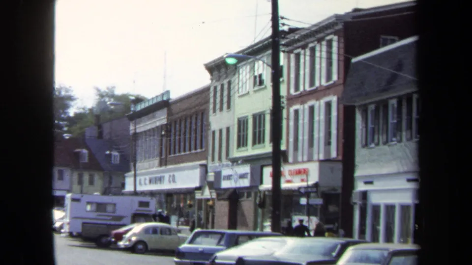 1970 Washington, DC street view with storefront signs, parked older cars, a utility pole, and small distant pedestrians along the sidewalk.