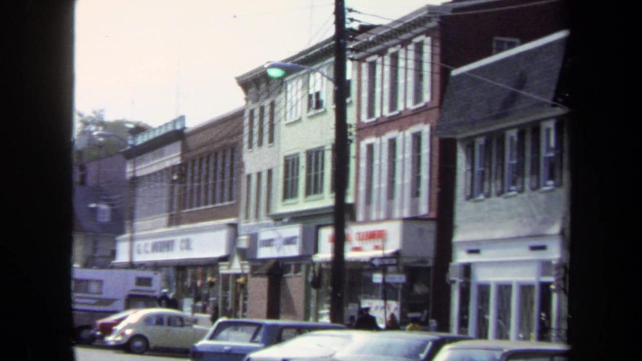 1970 Washington DC home-movie frame showing storefront buildings, parked cars, utility poles, overhead wires, and small pedestrians on the sidewalk.