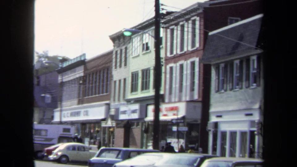 1970 Washington DC home-movie frame showing storefront buildings, parked cars, utility poles, overhead wires, and small pedestrians on the sidewalk.