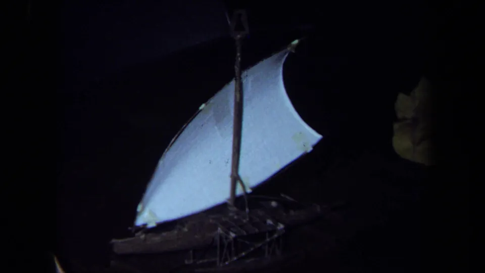 1971 California 8mm frame showing a small model sailboat with a white sail, mast, rigging, and dark hull surrounded by low light.