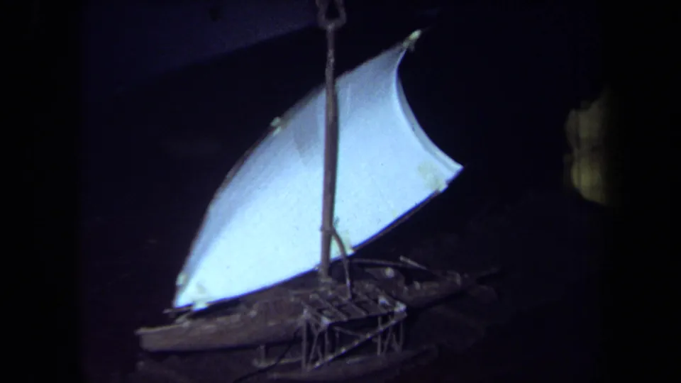 1971 California 8mm frame showing the small sailboat model from closer range, with a bright white sail, mast, rigging, and shadowed hull.