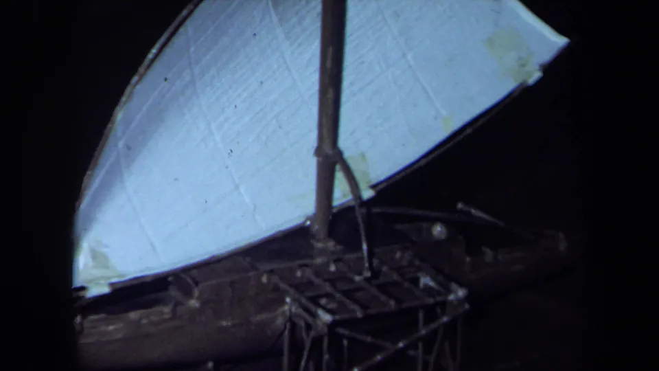 1971 California 8mm close-up of a model sailboat, emphasizing the white sail fabric, central mast, rigging, and dark wooden hull.