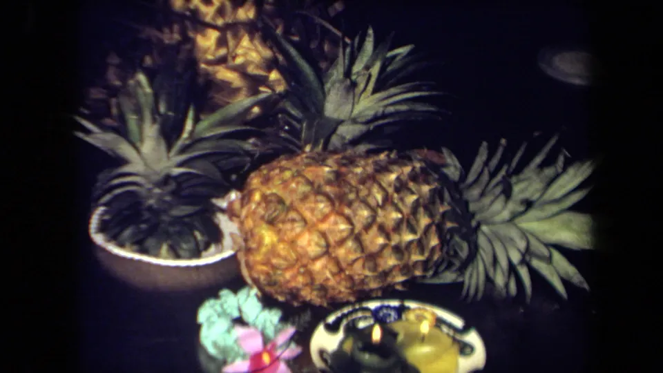 1971 California 8mm frame showing a pineapple, leafy garnish, pink flower, and two lit candles arranged as a tropical centerpiece.