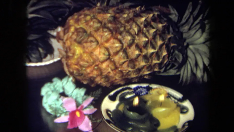Close 1971 California 8mm view of the tropical tabletop arrangement with pineapple, pink flower, green leaves, and two candles.