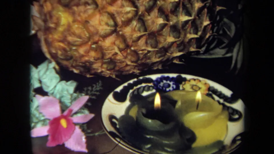 Tight 1971 California 8mm frame of a pineapple centerpiece with candlelight, decorative dish, green leaves, and a pink flower.