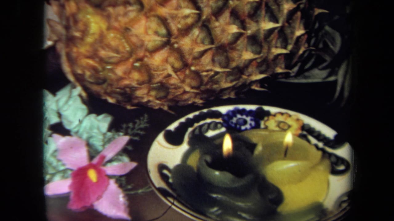 1971 California 8mm frame showing a pineapple, pink flower, patterned plate, and lit green and yellow candles.