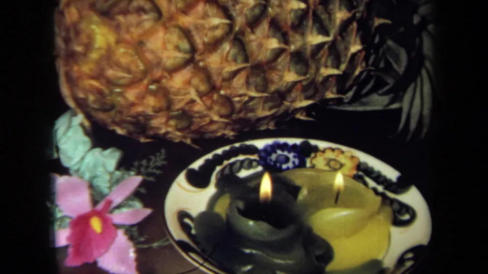 1971 California 8mm frame with pineapple, lit candles on a decorative plate, and a pink flower in a dark tabletop setting.