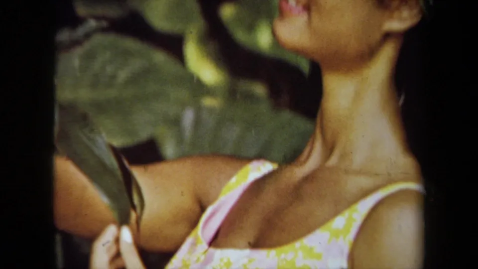 1971 California 8mm frame showing a woman in a yellow and pink floral dress among large green leaves, with her face mostly cropped.