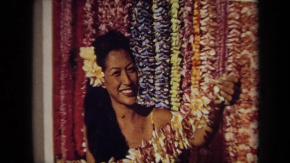 Close 1971 home-movie view of a woman pictured with a yellow flower in her hair and lei garlands behind her.
