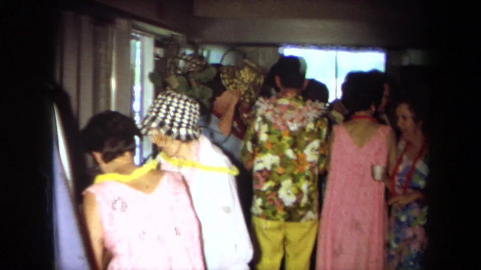 1971 California home-movie frame of party guests indoors, several wearing floral shirts, dresses, leis, and a checkered hat.