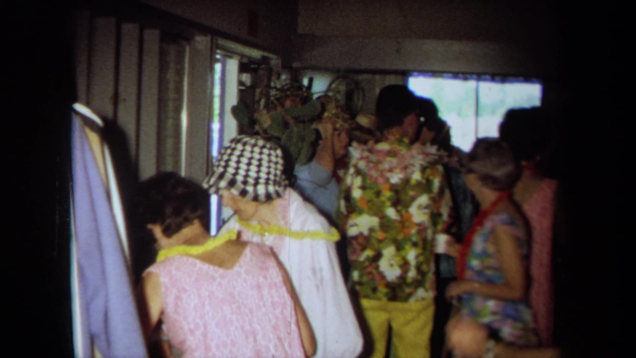 1971 California home-movie frame showing adults in leis, floral clothing, and a checkered hat inside a crowded private room.