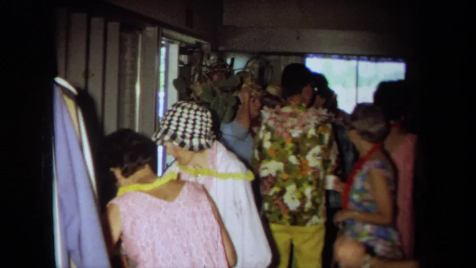 1971 California home-movie frame showing adults in leis, floral clothing, and a checkered hat inside a crowded private room.