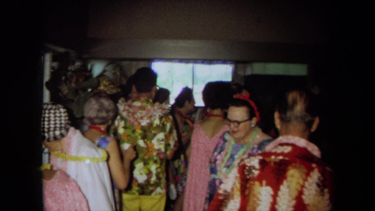 Crowded indoor party scene with adults wearing leis, floral shirts, and eyeglasses.