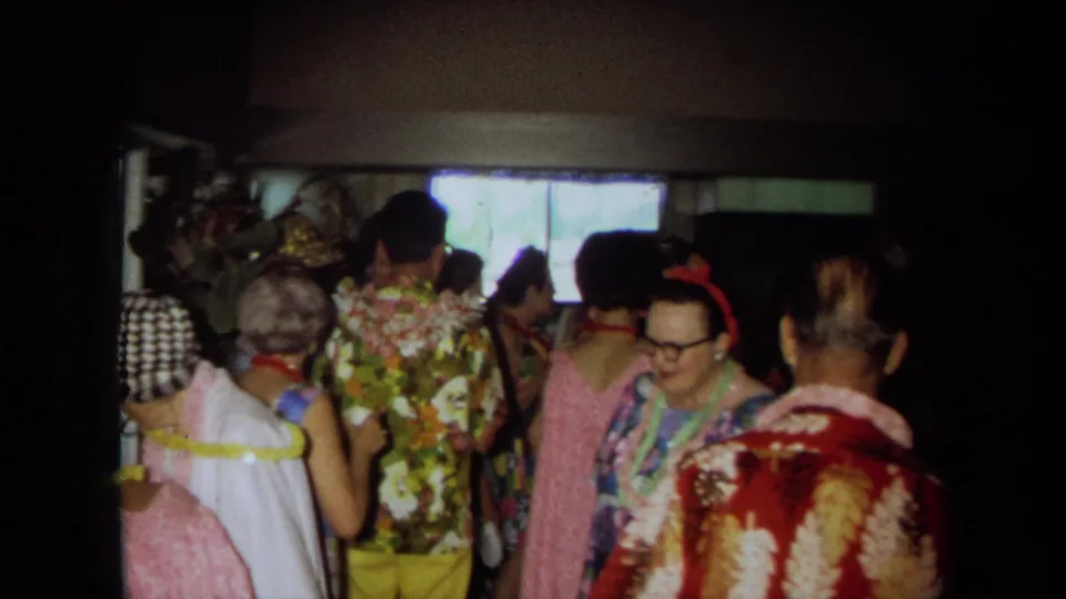 1971 California frame of adults socializing in a crowded room, including floral shirts, leis, eyeglasses, and a bright window behind them.