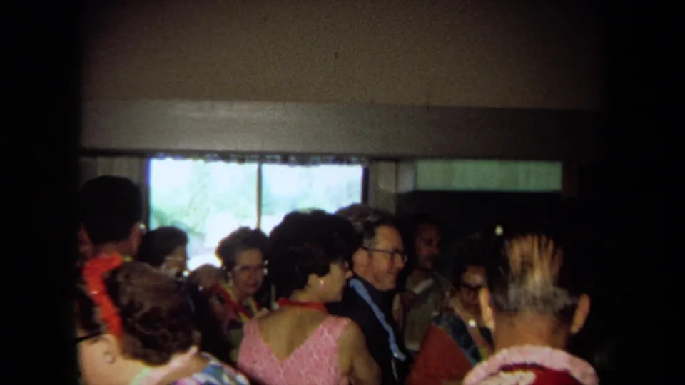 1971 California home-movie frame showing adults in leis and patterned party clothes talking in a dim private interior.