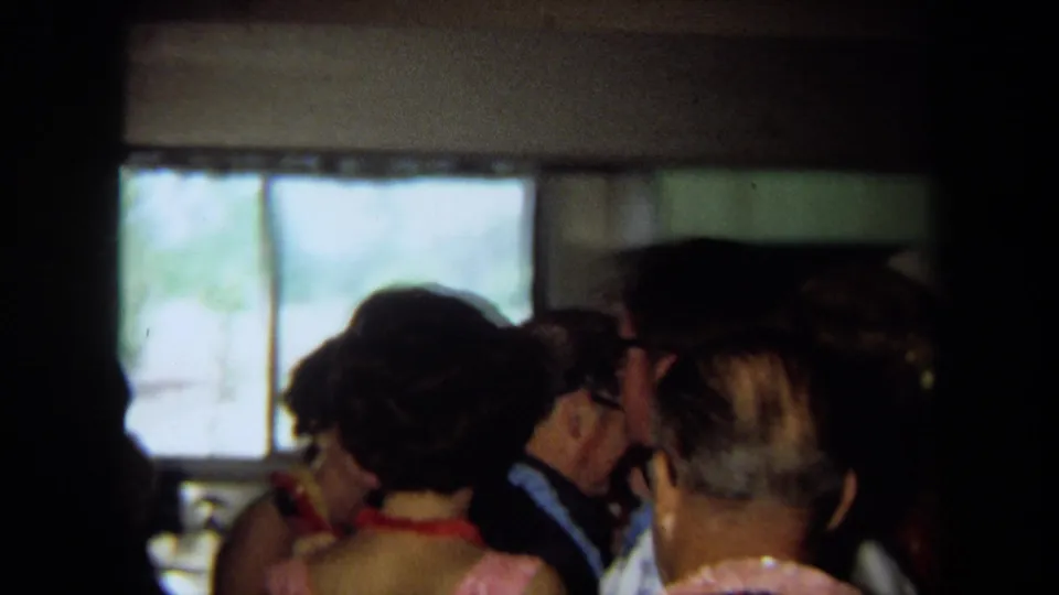 1971 California frame showing a backlit group of adults in a crowded private room, with leis and window light visible.