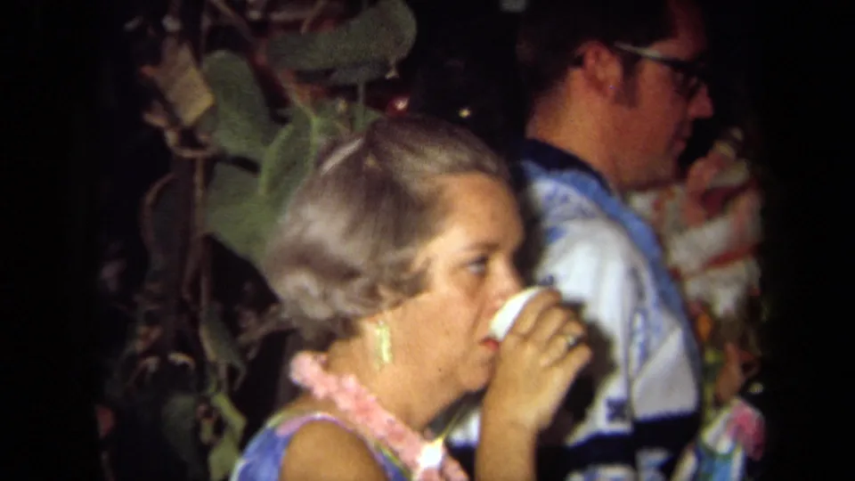 1971 California party frame with a woman in a pink lei sipping from a white cup near guests in aloha-style shirts.