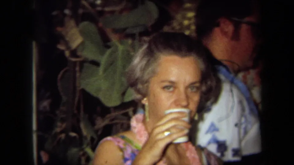 1971 California home-movie frame of a woman in a lei looking toward the camera while drinking at a Hawaii-themed party.