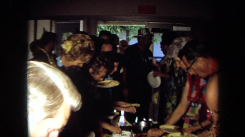 1971 California home-movie frame showing guests gathered around a serving table in a crowded Hawaii-themed interior.