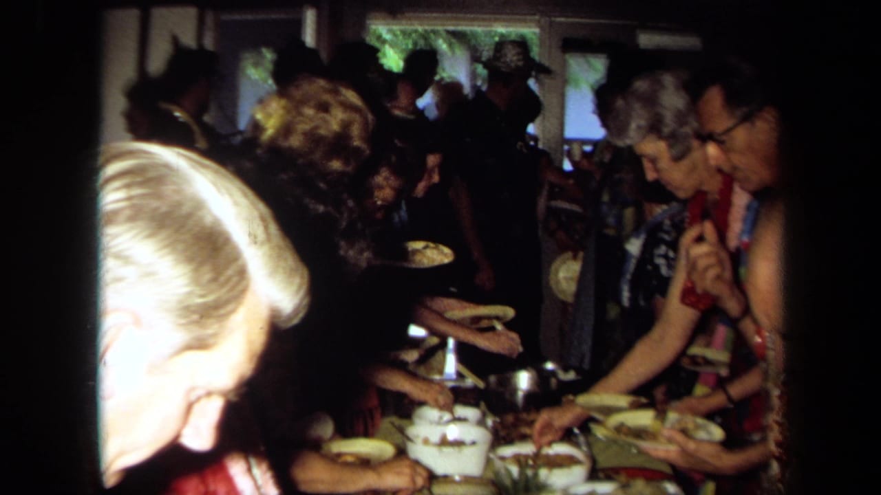 Guests in leis serve food from a crowded buffet table in a private room.
