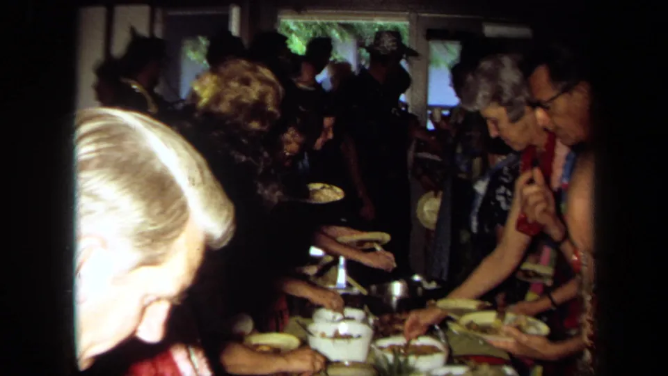 1971 California home-movie frame of adults in leis and patterned clothing serving food onto paper plates at a crowded buffet table.