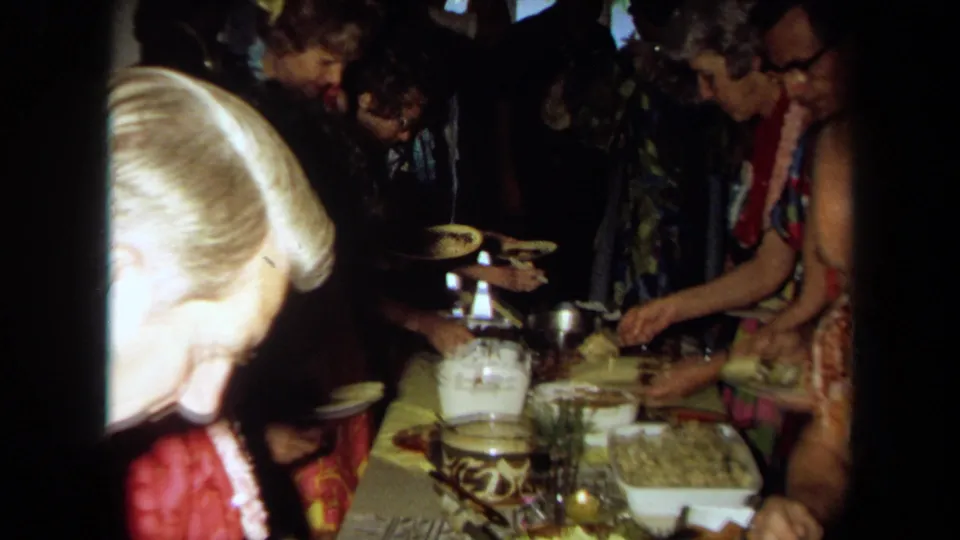 Adults in leis and aloha-style clothing gather around a long buffet table with bowls, plates, and serving dishes in a private California interior.