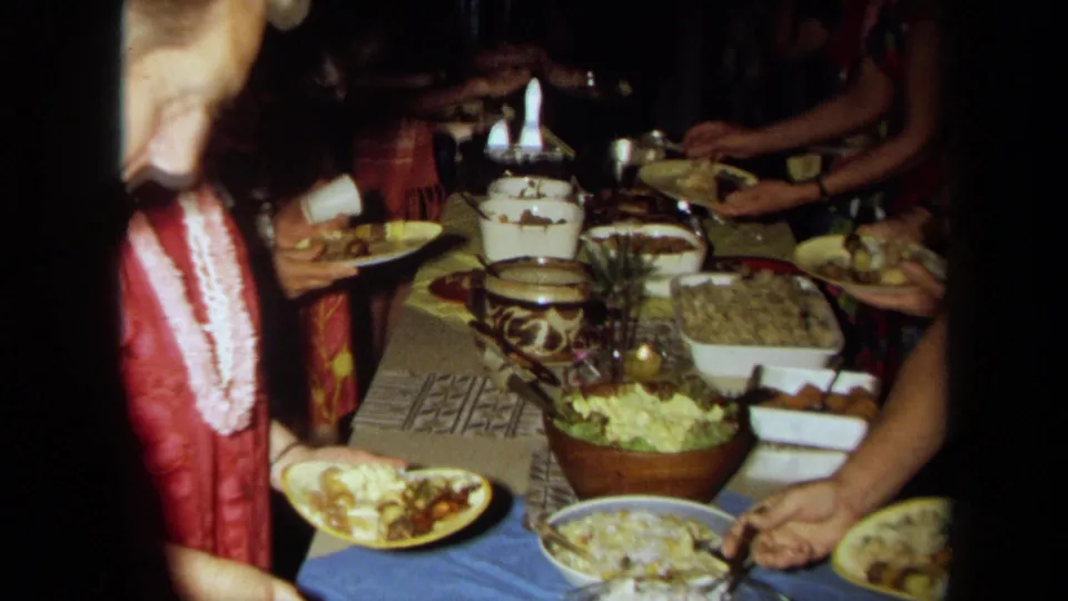 Close 8mm frame of a woman in red with a lei holding a paper plate beside rice, salad, bowls, and serving utensils at a party buffet.
