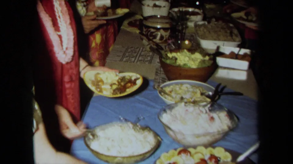 1971 California home-movie view of a buffet table with rice, salad, lemon slices, bowls, and adults in leis serving themselves.