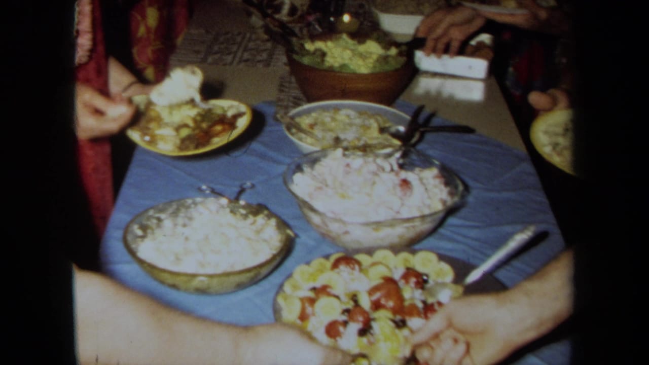 1971 California home-movie frame showing a blue-covered buffet table with salads, fruit, plates, and guests serving themselves indoors.