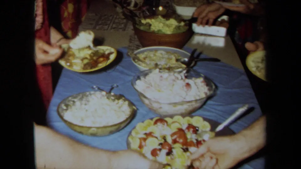 1971 California home-movie frame showing a blue-covered buffet table with salads, fruit, plates, and guests serving themselves indoors.
