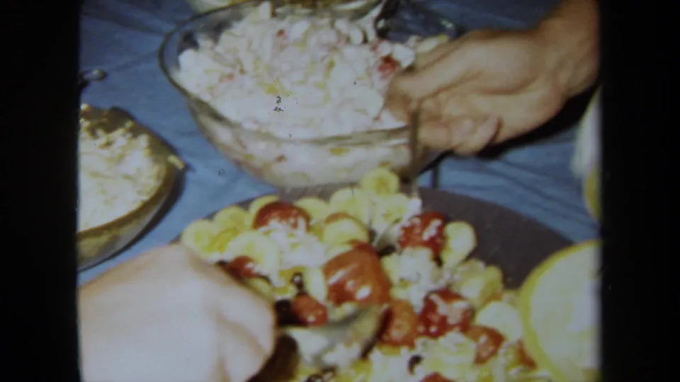 Hands serve food from a crowded indoor buffet table with salad bowls and fruit in this 1971 California home-movie frame.