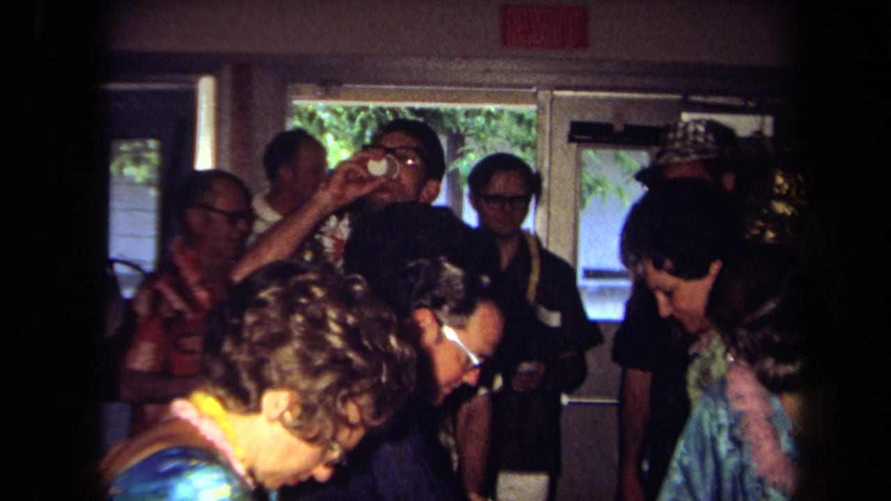 1971 California home-movie frame showing adults in leis and colorful shirts standing around a table in a crowded indoor party setting.