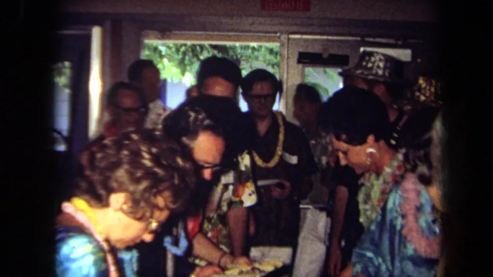 Adults at a 1971 California indoor party cluster around food and drinks, with leis, patterned shirts, doorway windows, and an EXIT sign visible.