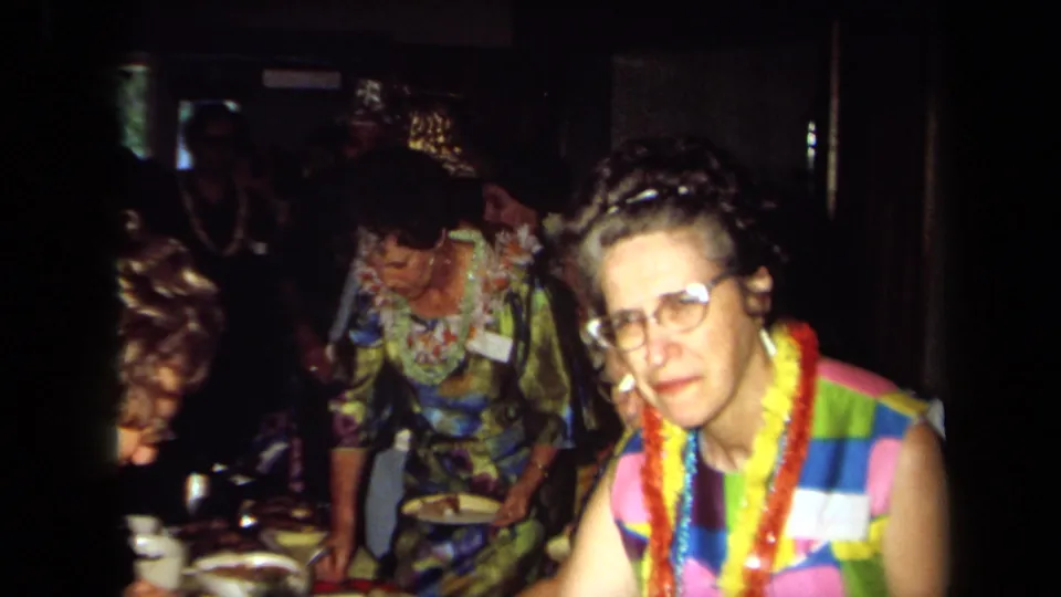 In this 1971 California party frame, a woman with a bright lei faces the camera as other adults gather at a buffet table behind her.